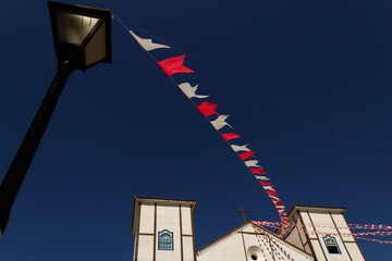 Um cordão com bandeirolas indo de um poste até a Igreja Matriz com céu azul ao fundo.