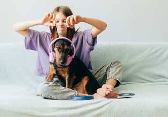 Happy teenage girl in lilac t-shirt sits on the sofa, listens to music has fun playing with the dog