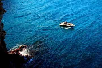 Breathtaking view from a high rocky shore of Polignano a Mare at a small motor boat with some tourists crossing deep wavy waters of the Adriatic sea