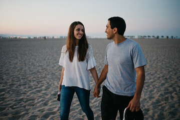 A Hispanic man with his smiling girlfriend are strolling on a sandy beach near the Balearic sea in Spain in the evening. A happy couple of tourists on a date at twilight in Valencia.