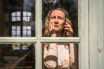 Sad woman talking on phone at home and crying while standing near the window