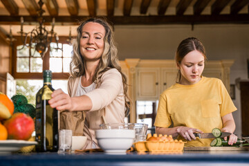 Smiling adult woman taking oil from the table while her serious girl chopping vegetables