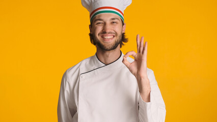 Young successful chef dressed in uniform showing okay sign at camera over yellow background....