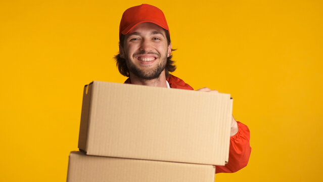 Beautiful delivery man wearing uniform holding cardboard boxes over colorful background. Male bearded courier enjoying his work standing with parcel boxes in studio