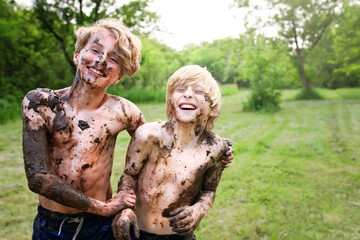 Two happy little kids are laughing while outside covered in mud by the river on a summer day.