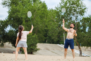 Back view of little girl with long dark hair wearing white T-shirt, grey skirt, playing badminton with middle-aged bearded shirtless man in shorts on sandy beach of river. Summer activities. Family.