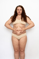 Portrait of pouty overweight woman with long curly hair wearing beige underwear, squeezing excess...