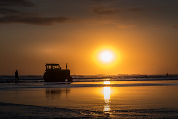 Fototapeta premium Silhouette of a tractor on the beach at sunset over the sea and reflection on the water