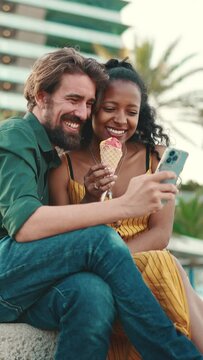 VERTICAL VIDEO: Close up of happy man and smiling woman watching video on smartphone. Close-up of a joyful young interracial couple browsing photos on mobile phone on urban city background