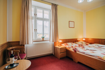 Old hotel room with two individual beds pushed together. Room has standard equipment in form of lamps, bedside tables and a seating area. Room also has window and yellow-painted walls with wood trim.