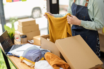 Portrait of a small startup Asian female entrepreneur SME owner picking up a yellow shirt before packing it in an inner box with a customer. Online Business Ideas and Freelance