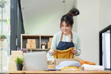 Portrait of a small startup Asian female entrepreneur SME owner picking up a yellow shirt before packing it in an inner box with a customer. Online Business Ideas and Freelance