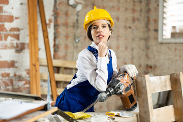 Confident female contractor in blue overalls and yellow safety hard hat standing at construction...