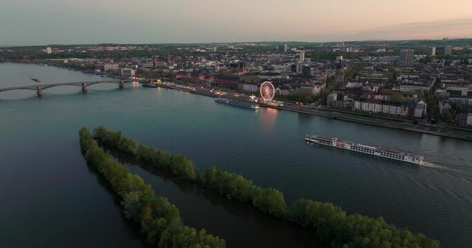 Mainz summer night with city lights and ships on the Rhein river in front of orange sky by a cinematic drone shot