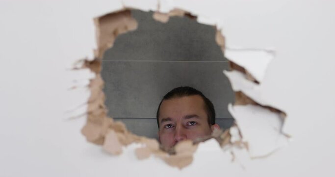 Young man raises his head and sees that someone is watching him through a hole punched in plasterboard door, so he makes a dumbfounded look and says something, front view