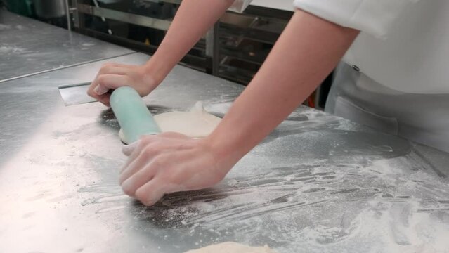 Close up of chef's hand in white cook uniforms with aprons are kneading pastry dough with roller, preparing bread, pies, and fresh bakery food, baking in oven at stainless steel kitchen of restaurant.