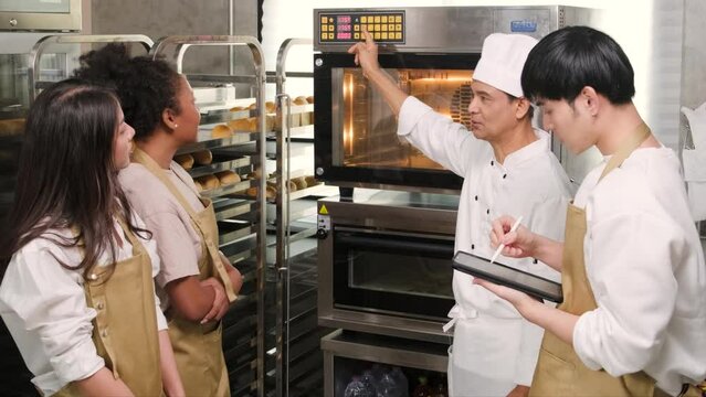 Senior Asian male chef of culinary learning course teaches and explains about electric oven to cooking class students for baking pastry dough for bread and bakery foods in stainless steel kitchen.