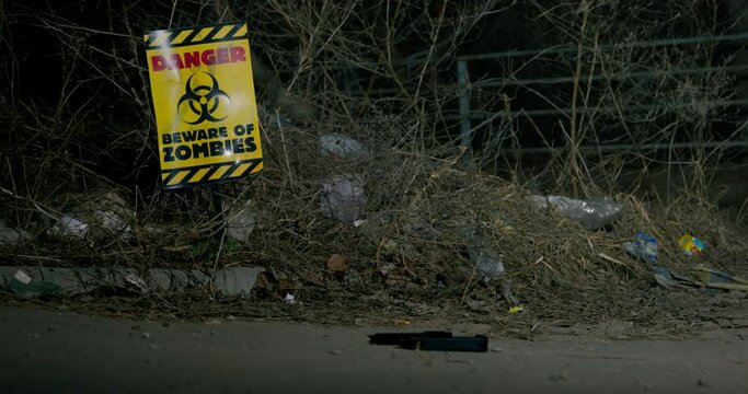 Person in rubber boots throws guns on the ground and walks away, he has run out of ammo, low-angle view. There is a sign in ground warning of danger of zombie attack