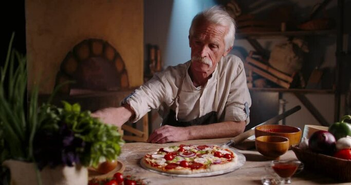 Senior experienced chef is preparing pizza for serving, sprinkling basil leaves. Old cooker using traditional recipe in pizzeria, looking at camera 4k portrait footage