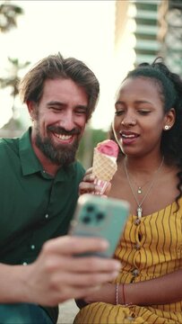 VERTICAL VIDEO: Close up portrait of happy man and smiling woman watching video on smartphone. Close-up of joyful young interracial couple browsing photos on mobile phone on urban city background