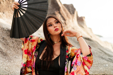 Fashion photo of elegant asian woman in silk kimono  holding fan and posing over rocks on the beach.