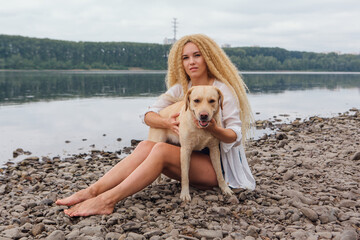 Fototapeta na wymiar Young beautiful woman with blond curly hair sitting with her labrador retriever dog on the river shore