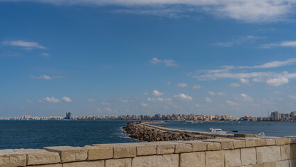 View of the Mediterranean Sea from the embankment of Alexandria. The breakwater is visible, city buildings in the distance. Blue sky with clouds. Egypt.