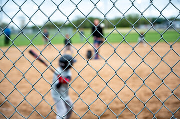 Selective focus on chain link fence with a youth baseball game defocused and blurred in the background - obrazy, fototapety, plakaty