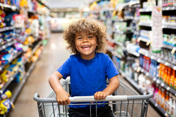 Funny little boy laughing while sitting in a shopping cart during a family trip to the grocery store. He is excited to buy something new	 - obrazy, fototapety, plakaty