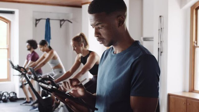 Young adult black male typing on electronic tablet at an indoor fitness gym. Confident fitness professional providing guidance.