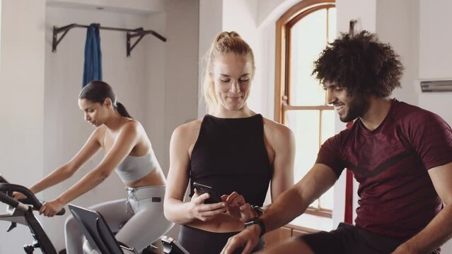 Caucasian female chatting to middle eastern male while cycling on exercise bike at an indoor fitness gym. Showing him cellphone image.