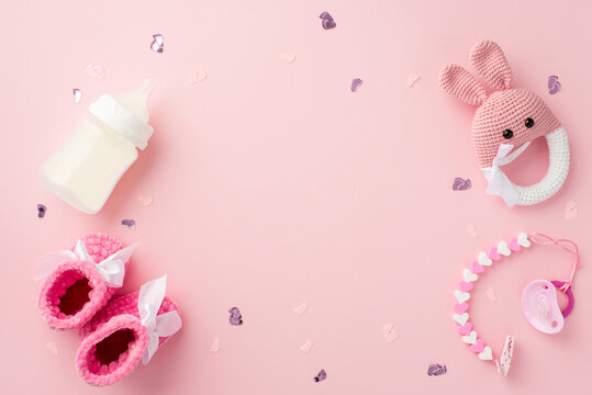 Baby accessories concept. Top view photo of tiny pink shoes pacifier chain bottle knitted bunny rattle toy and confetti on isolated pastel pink background with copyspace in the middle