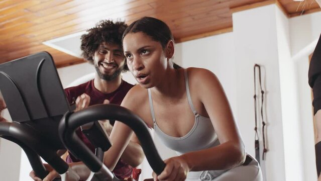 Diverse group of friends cheering biracial female cycling on stationary bike at an indoor fitness gym