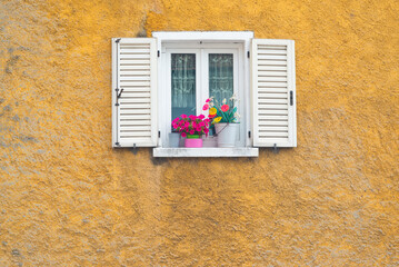 White wooden window opened on a yellow wall with colorful flowers in the ancient village named "Argegno", Lake Como, Italy.