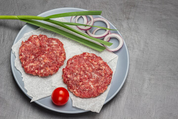 Two raw beef burgers with green onion and tomato on gray plate.
