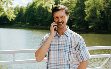 Smiling handsome man talking on smartphone while standing on a bridge in a park on a sunny day, copy space. Happy caucasian guy using mobile phone and looking away outdoors.