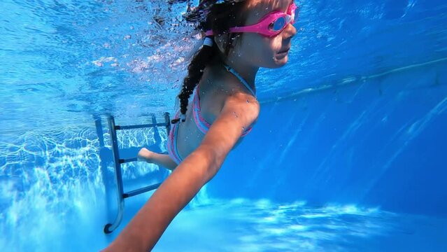 Little girl study to swim underwater in pool. Child in blue bikini and pink glasses holds toy and moves forward. Having fun in pool. Sport good for healthy. Concept vacation and hobby
