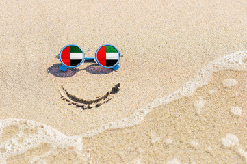 Fototapeta na wymiar A painted smile on the beach and sunglasses with the flag of the United Arab Emirates. The concept of a positive and successful holiday in the resort of the United Arab Emirates.