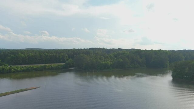 aerial footage of the rippling waters of Lake Acworth surrounded by lush green trees, grass and plants with cars driving on the road and blue sky with clouds at Proctor Landing Park in Acworth Georgia