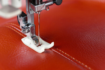 Modern sewing machine presser foot with a needle sews red leather. Sewing process of decorative seam on leatherette with special leather presser foot. Close up, copy space.