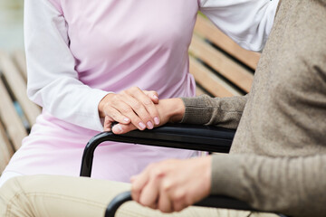 Close-up of unrecognizable woman in uniform sitting on bench and touching hand of patient in wheelchair while giving care to him