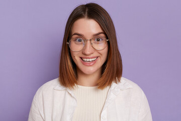 Indoor shot of attractive young woman wearing white clothing and glasses posing isolated over purple background, looking at camera with happy positive expression.