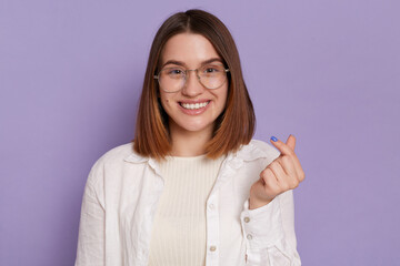 Pretty dark haired woman with gentle smile makes korean like sign expressing love to you, wearing white shirt, posing isolated over purple background. Miniheart gesture.