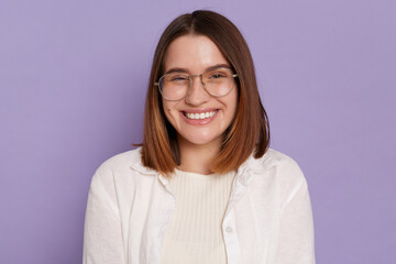 Portrait of delighted satisfied young woman wearing white clothing and glasses posing isolated over purple background, looking at camera with toothy smile, expressing happiness.