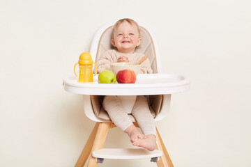 Portrait of extremely satisfied toddler baby girl dresses in beige jumper sitting in high chair and...