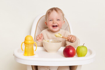 Image of deligted laughing infant baby girl dresses in beige jumper sitting in high chair and...