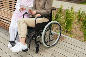 Obraz premium Unrecognizable nurse in uniform sitting on bench in yard of hospital and showing video on tablet to handicapped man in wheelchair
