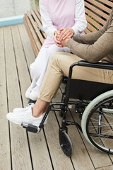 Unrecognizable qualified nurse in uniform sitting on bench outdoors and holding hands of disabled...