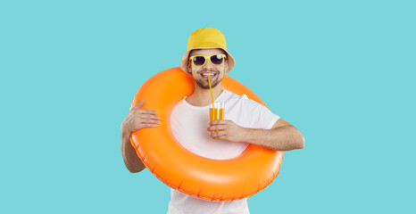 Happy young man with inflatable swimming circle and with glass of juice enjoys summer vacation. Cheerful funny guy in summer clothes drinks juice on light blue background. Summer relaxation concept. - obrazy, fototapety, plakaty