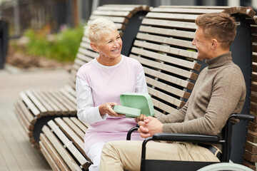 Jolly attractive mature lady with short hair sitting on bench outdoors and opening lunch box for...